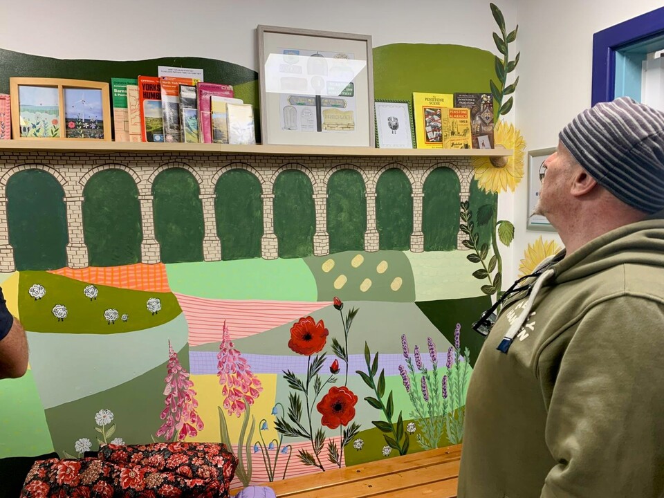 A person wearing a striped beanie and green hoodie looks at a colorful mural of a countryside scene with flowers, fields, and an arched stone bridge, with books and framed pictures displayed on a shelf above.